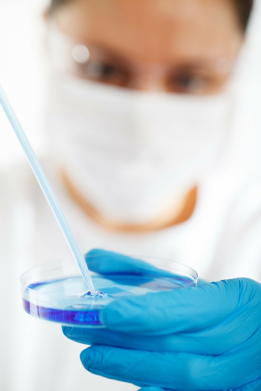 A masked scientist uses a pipette to draw up blue liquid from a petri dish.
